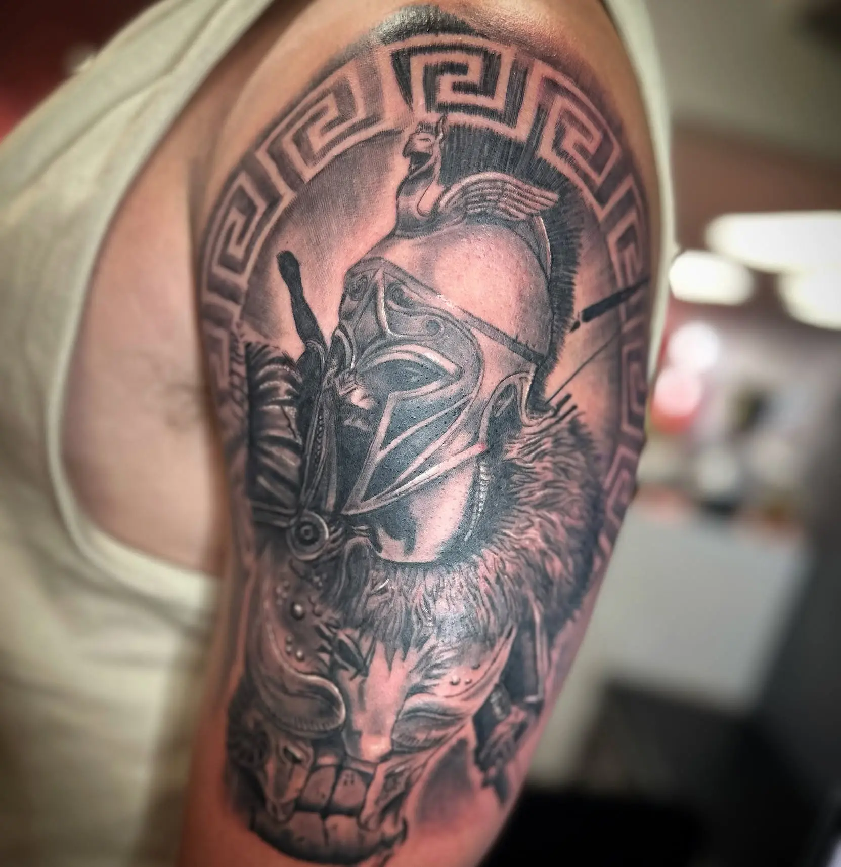 Black and grey Spartan warrior in a crested helmet on an upper arm, framed by a Greek key border